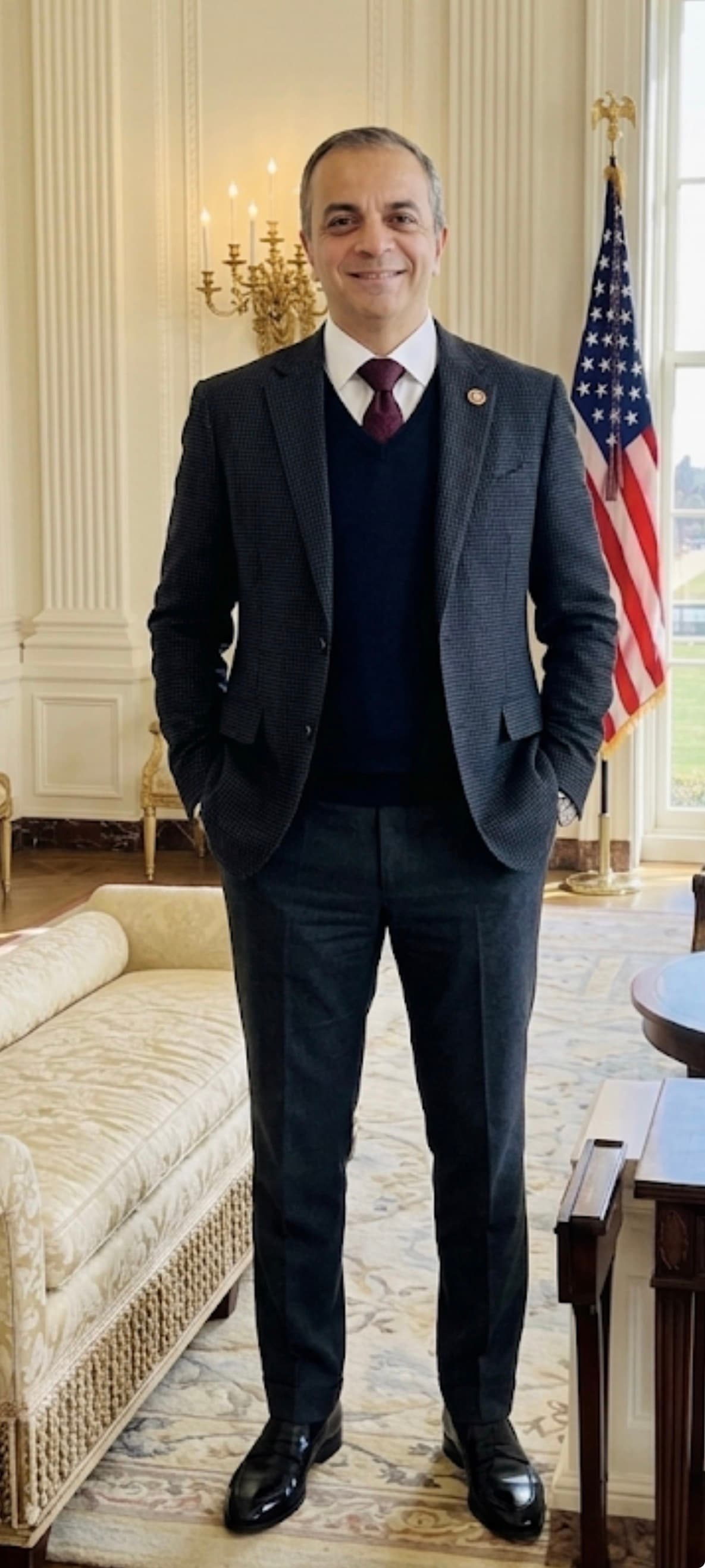 Smiling man in suit and sweater vest stands in a formal room with American flag.